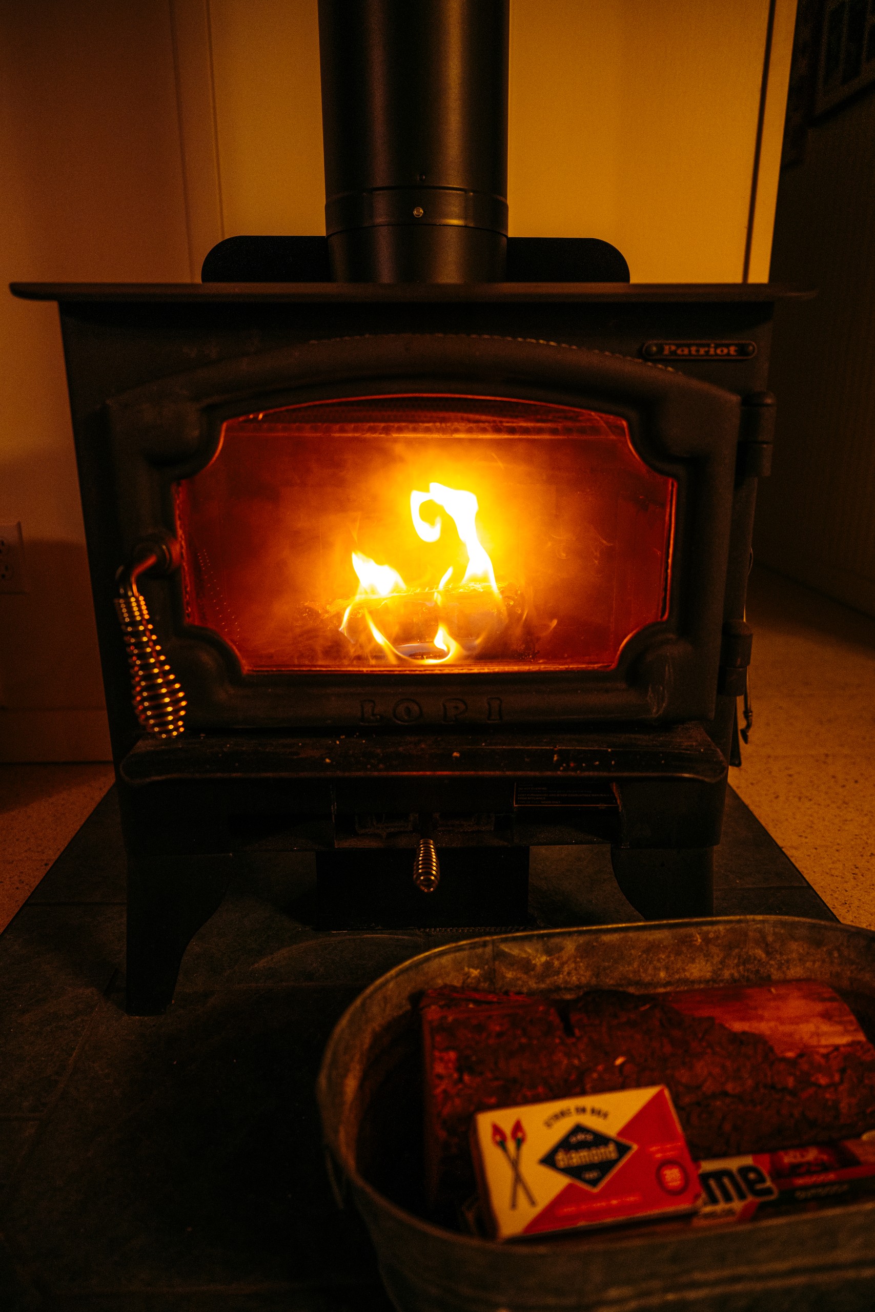A fire burning in the wood stove with a tin of firewood and matches beside it